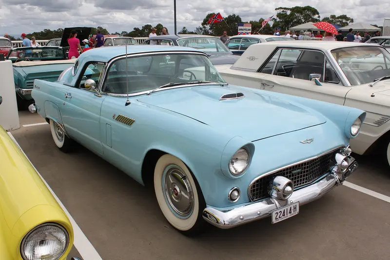 A pale blue 1956 Ford Thunderbird with a white hardtop parked at a car show